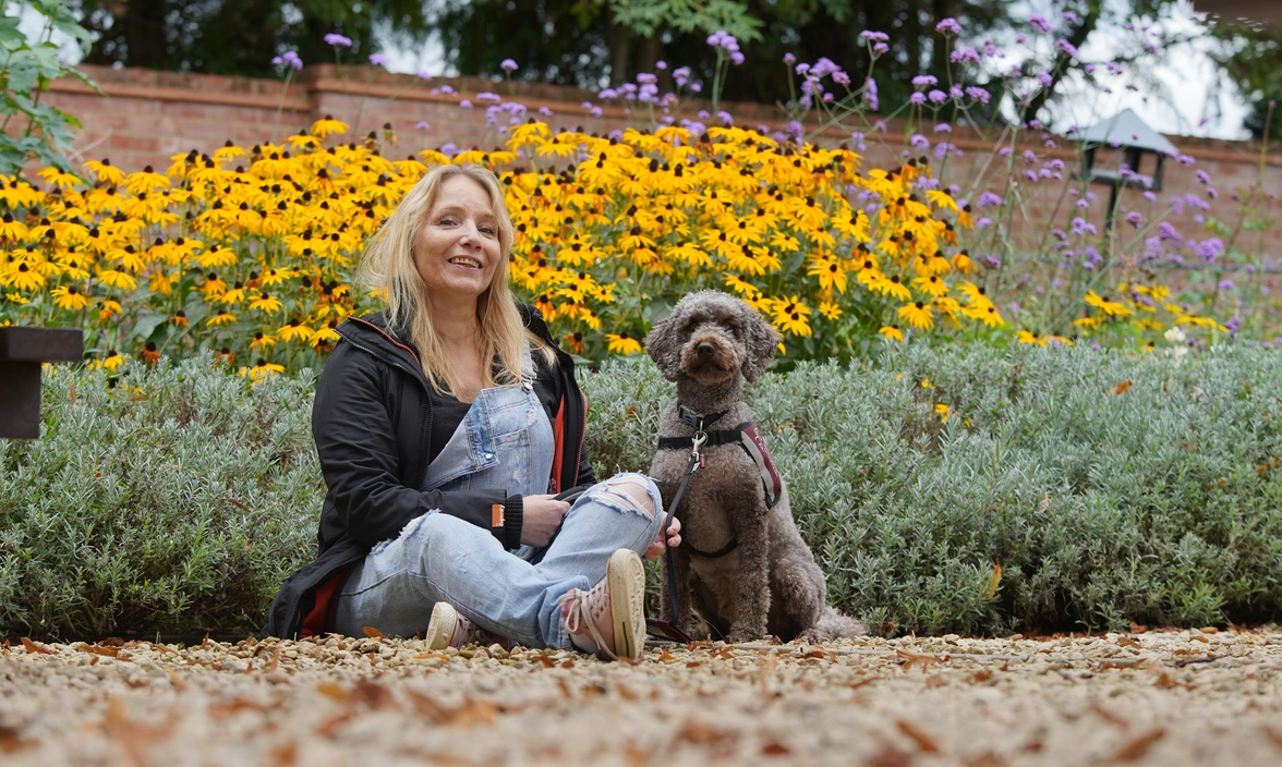 A blond woman and a dog with short, curly dark fur are sitting on the ground in front of many bright yellow flowers