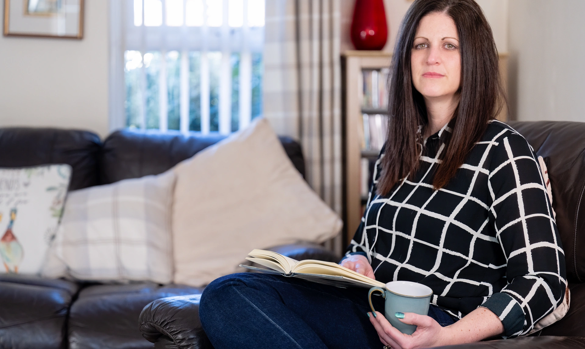 A woman with shoulder length dark hair sitting on a lounge chair with a book in her lap and a mug in hand. She looks solemn.