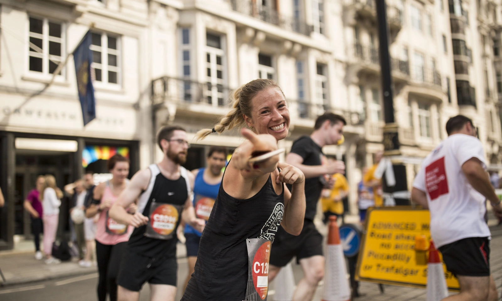 A female runner smiles broadly while pointing at the camera. Other runners can be seen in the background.