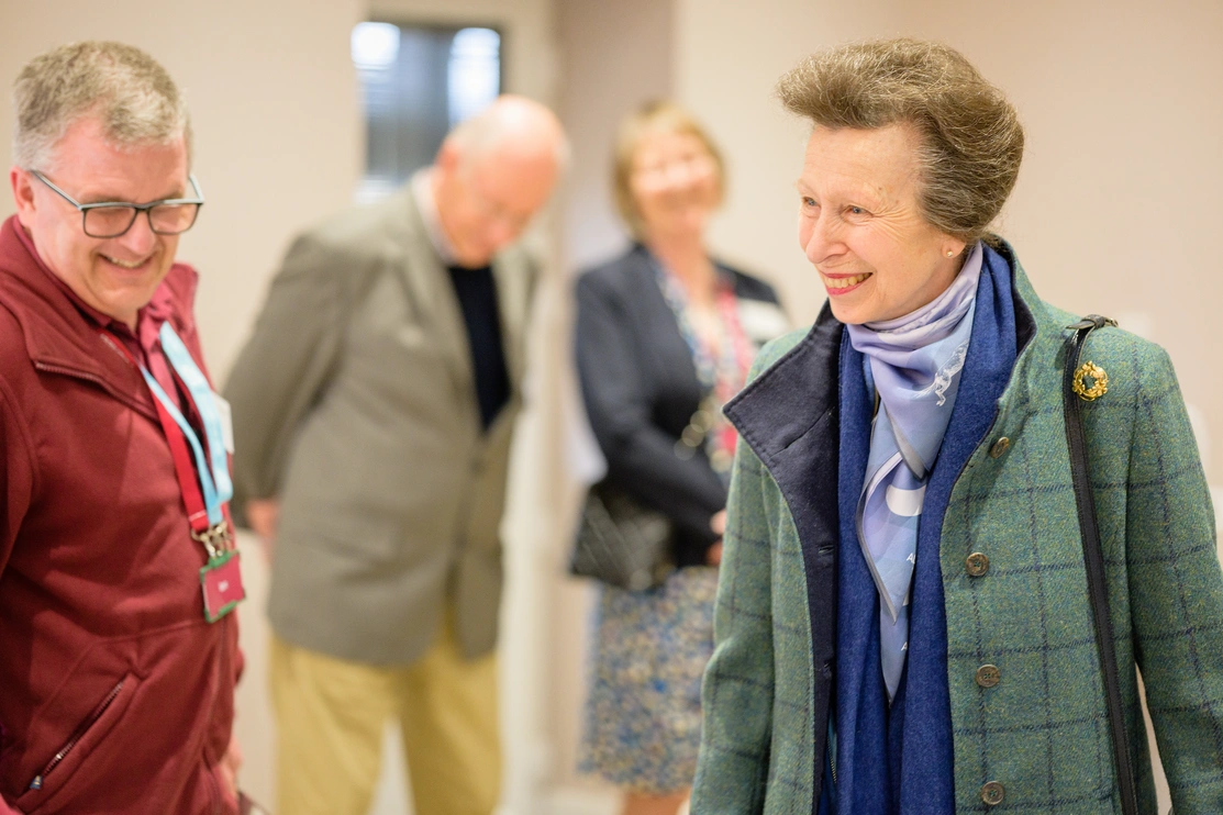 HRH The Princess Royal in a green coat and blue scarf and blouse looking to the left and smiling with a Hearing Dogs volunteer and some people in the background