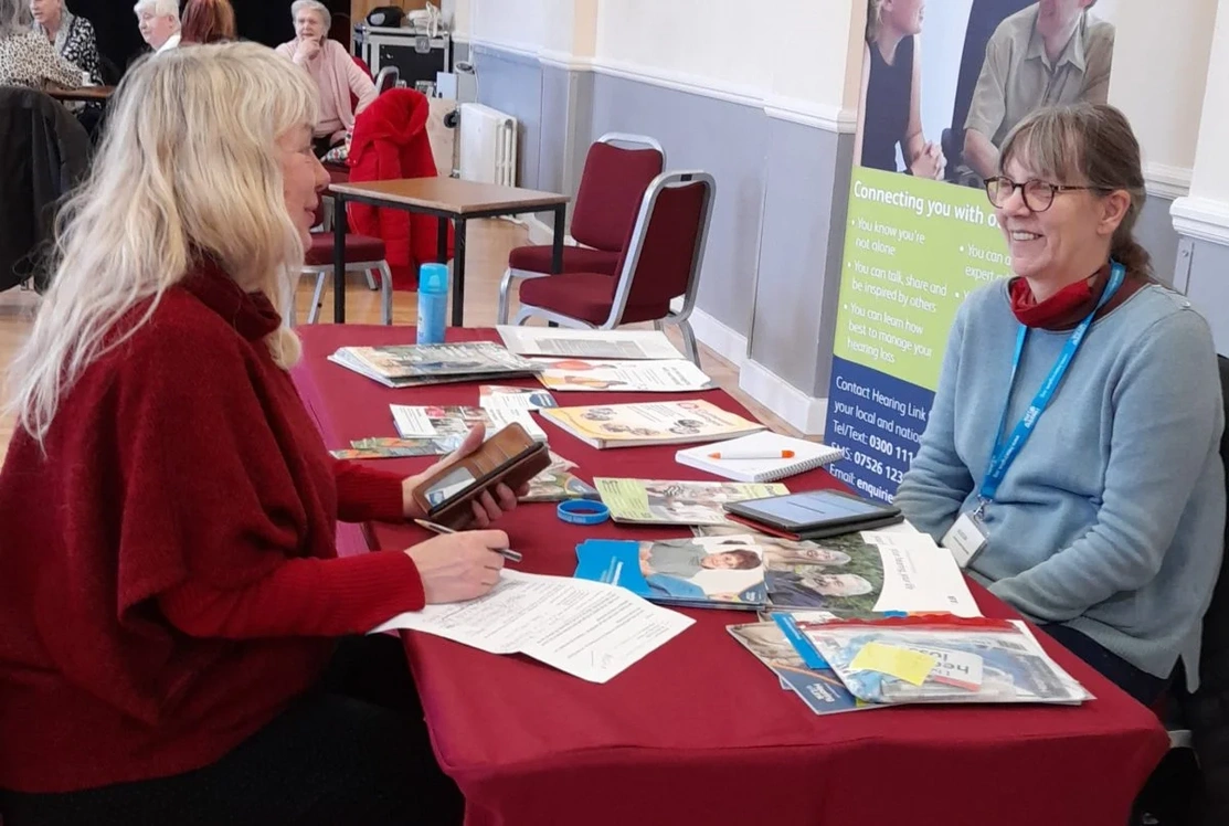 One woman talking to another one while sat at a table with literature on it