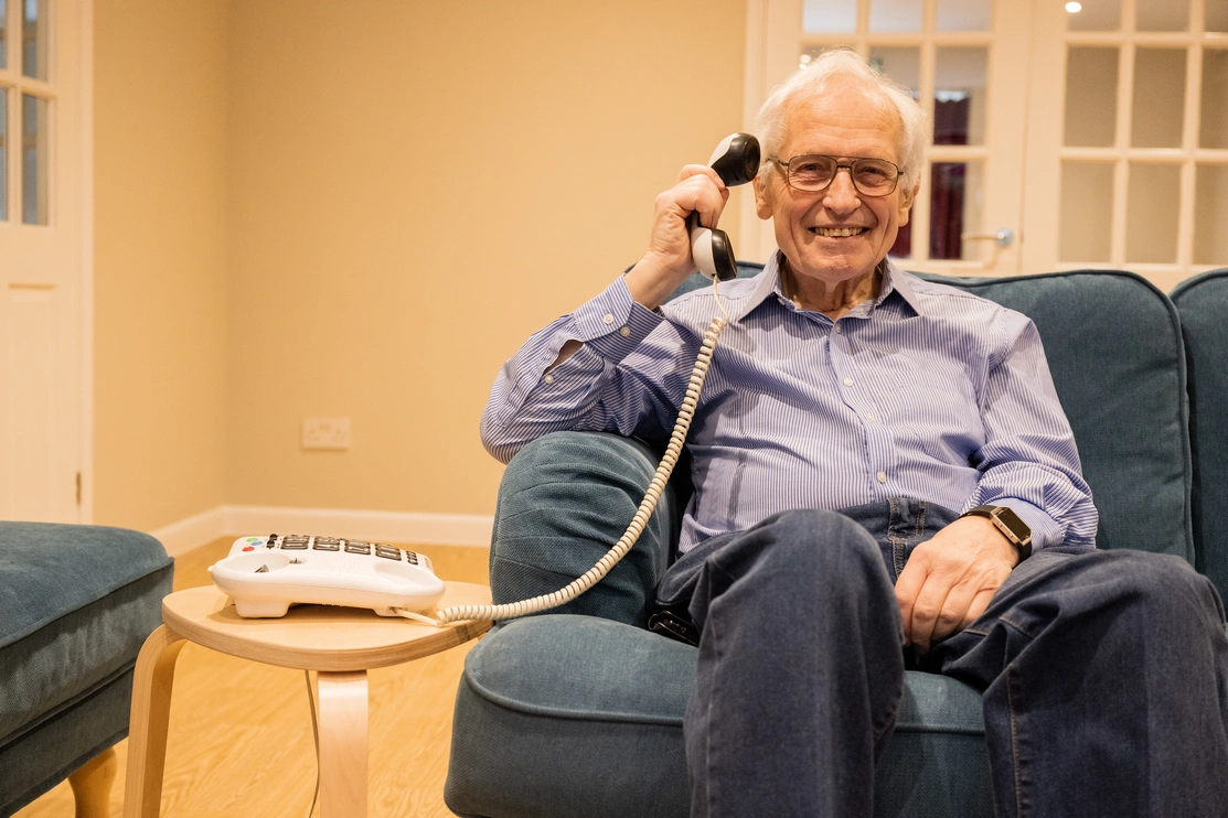 A man smiling sat on a sofa holding up a landline phone receiver to his ear