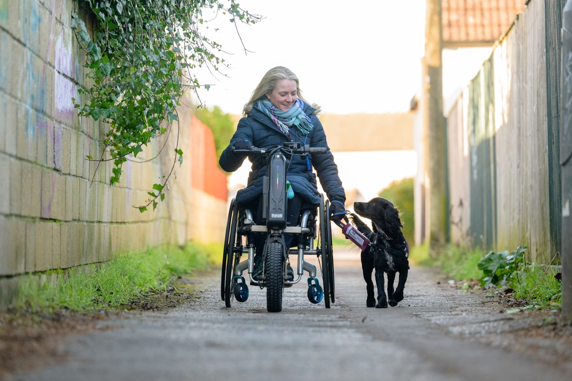 Woman in a wheelchair walking with a hearing dog on a lead down a path outside 