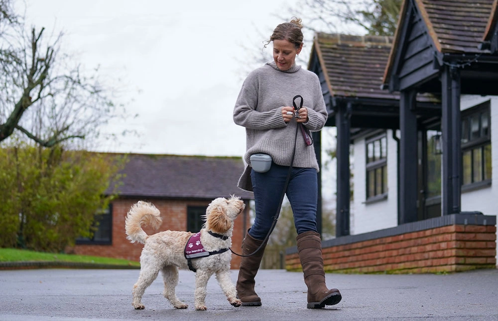 A woman walking a dog on a lead on a tarmac area with building in the background, the dog is looking attentively at the woman