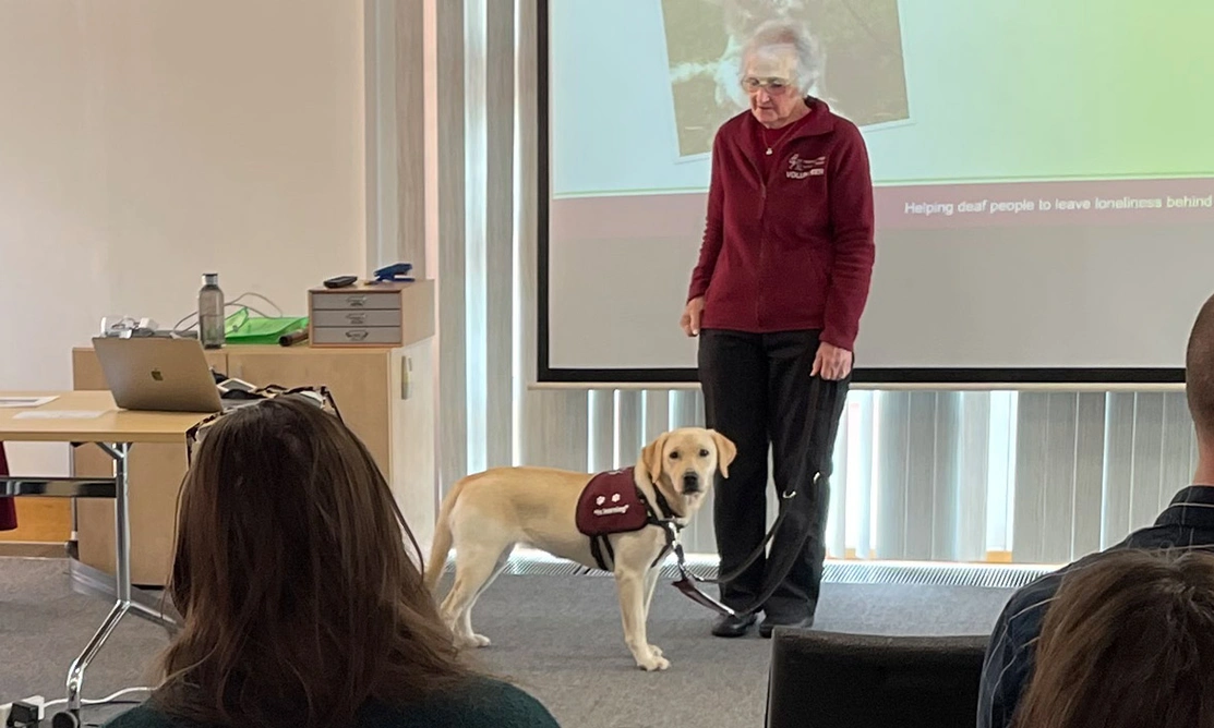 Lady standing in front of an audience, alongside a yellow Labrador wearing a Hearing Dogs training jacket