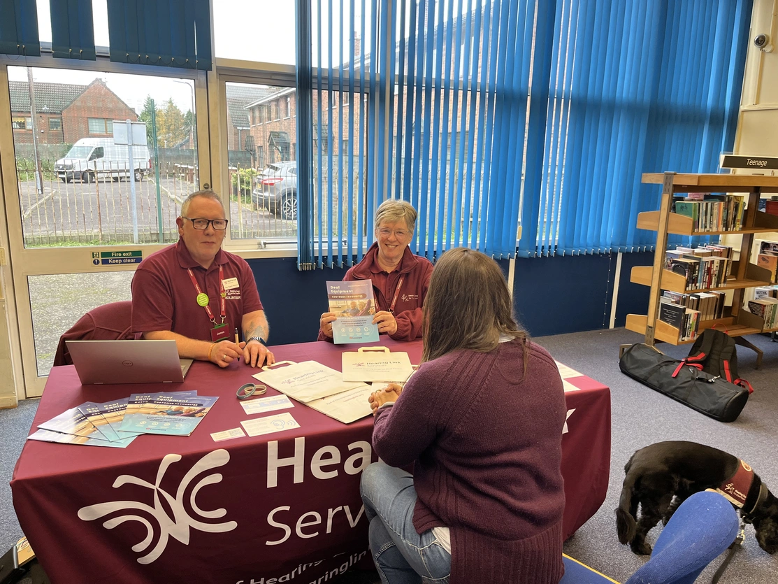 A man and two women sitting a table in a library, one woman has her back to camera. The table has various literature on it.
