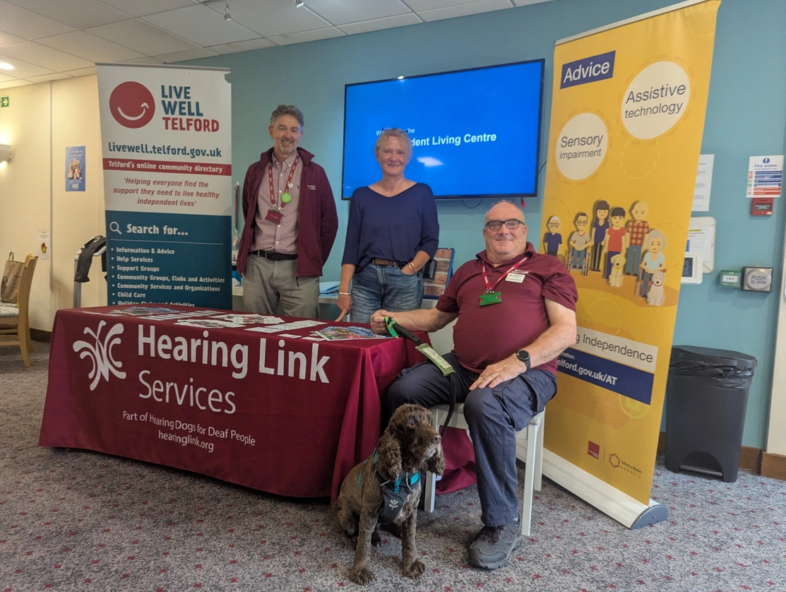 Three people and a dog assembled around a display table. A man and woman standing behind the table and a man sitting at the end holding the dog's lead