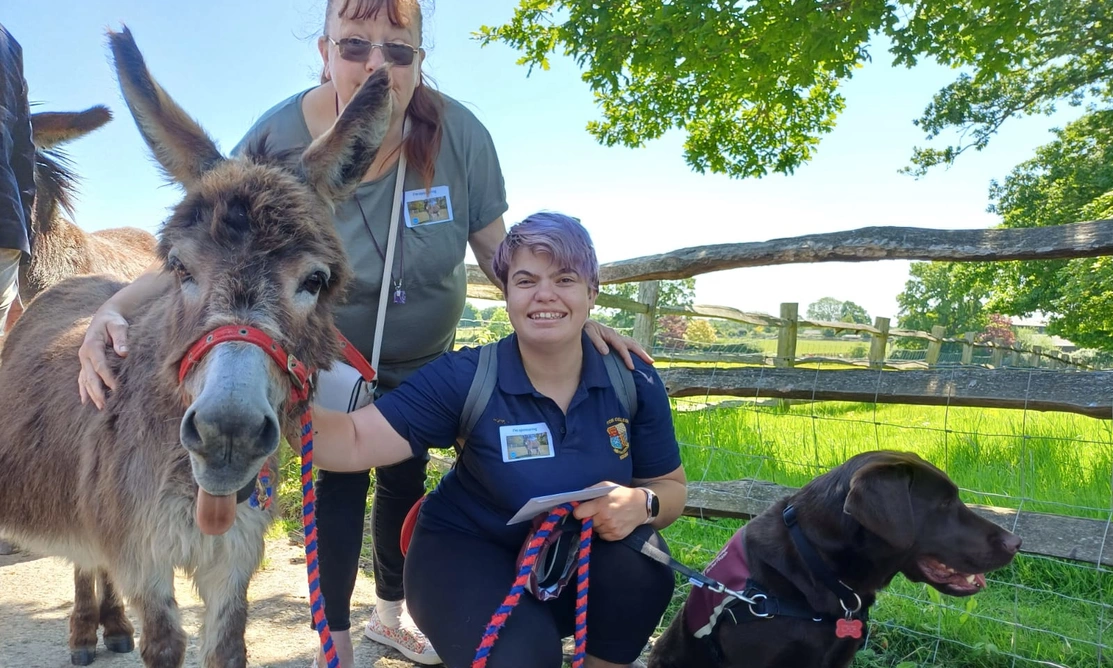 Two women are pictured with a donkey and a black labrador in an outdoor setting. One woman is standing, the other with short colourful hair is kneeling and holding the dog's leash.