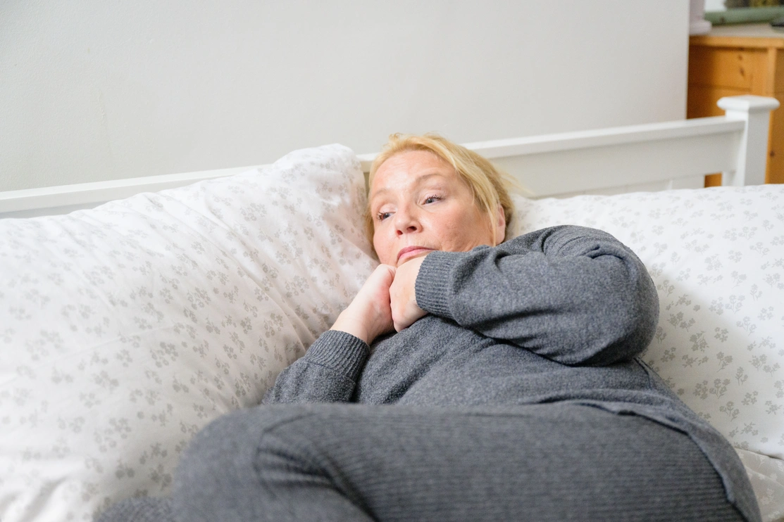 A woman in grey clothing lying on a bed. Her face shows worry and anxiety.