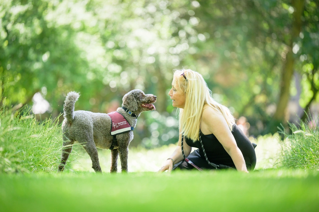 A blonde woman and a dog are in a grassy, tree filled location. She is sitting on the grass looking at the dog who is looking back at her, tail wagging.