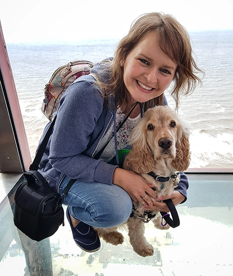 A smiling woman kneeling and hugging a light haired dog. They appear to be high above ground inside a structure with windowed walls and floor. A body of water can be seen behind them.
