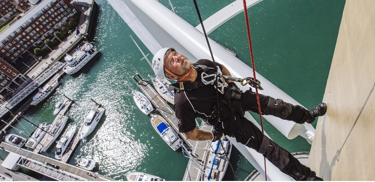 Image shows a man standing on the side of a building looking up. Water and boats can be seen far below him
