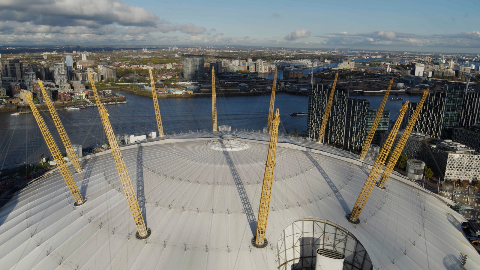 A aerial  view of the London O2 arena with its white dome and yellow pillars. The Thames river can be seen in the background.