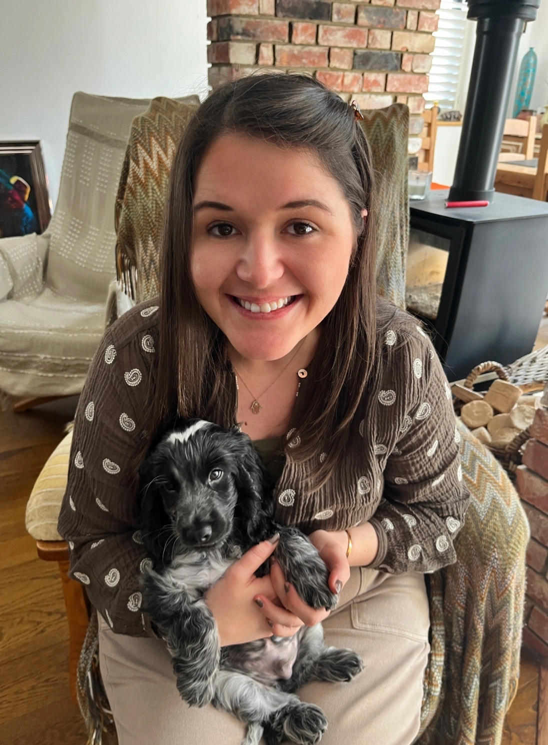 Jordan is sitting on a chair holcing a cute puppy. She has dark brown hair and is smiling.