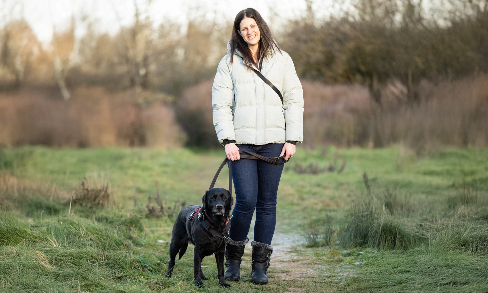 A woman wearing a white jacket standing in a field with a black dog at her side.