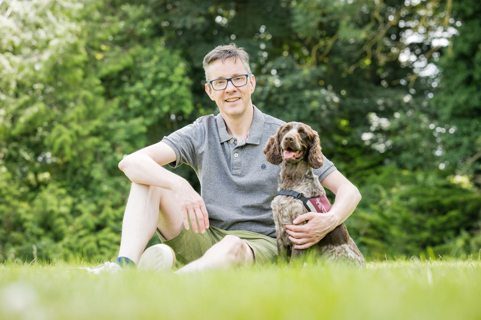 A smiling man, wearing spectacles, sitting on grass, with trees visible behind him. His left arm is around a brown and white cocker spaniel, which is wearing a hearing dogs jacket.