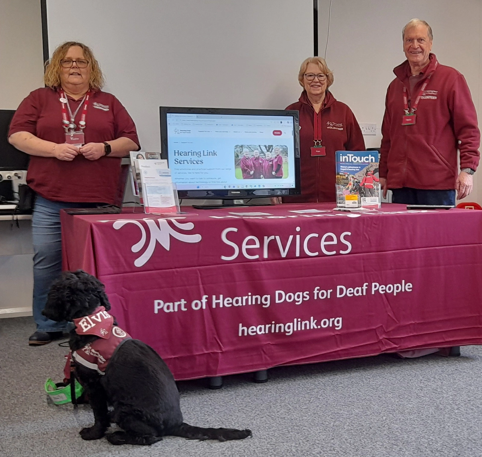 2 women and a man standing behind a table draped in a burgundy cloth. A black hearing dog sits in front.