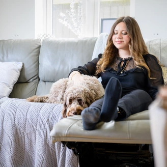 A young woman sat on a sofa smiling with her arm around a light brown cockapoo