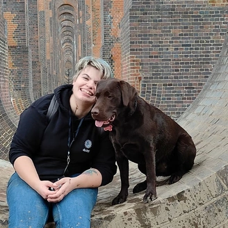 A smiling woman and a dark brown Labrador are sitting within a large brick edifice.
