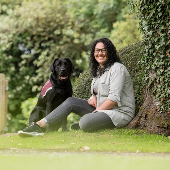 Alt Text: Smiling woman with long dark hair and casual clothes sits on some grass against a tree with a black labrador next to her wearing a burgundy Hearing Dogs jacket