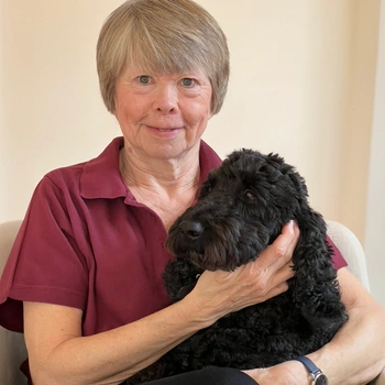 Jan with a black cockerpoo. Jan has short dark blond hair. She is wearing a burgendy Hearing Dogs polo shirt and has one hand holding the collar of the cockerpoo next to her.