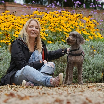 A woman sat on gravel with bright yellow flowers behind her and her hearing dog poodle stood next to her