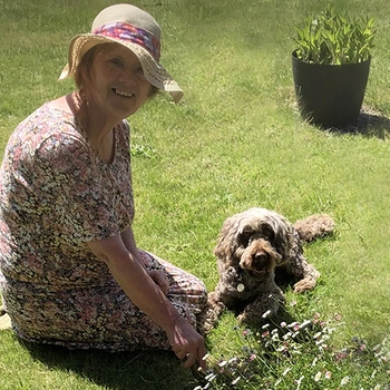 A smiling woman sat on grass looking happily at the camera with her sound support dog laying in the grass beside her