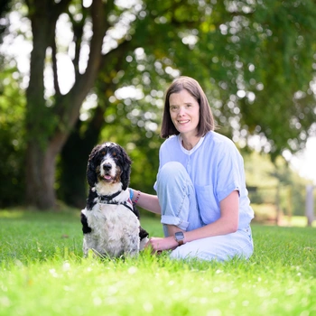 Young woman with brown hair and a blue top smiling and crouching next to a black, white and orange Spaniel outside on grass with trees in the background
