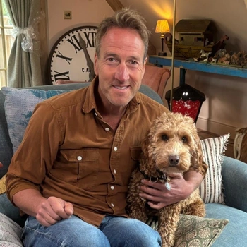 A man (ben fogle) sitting on a blue couch, his arm around a light coloured dog.
